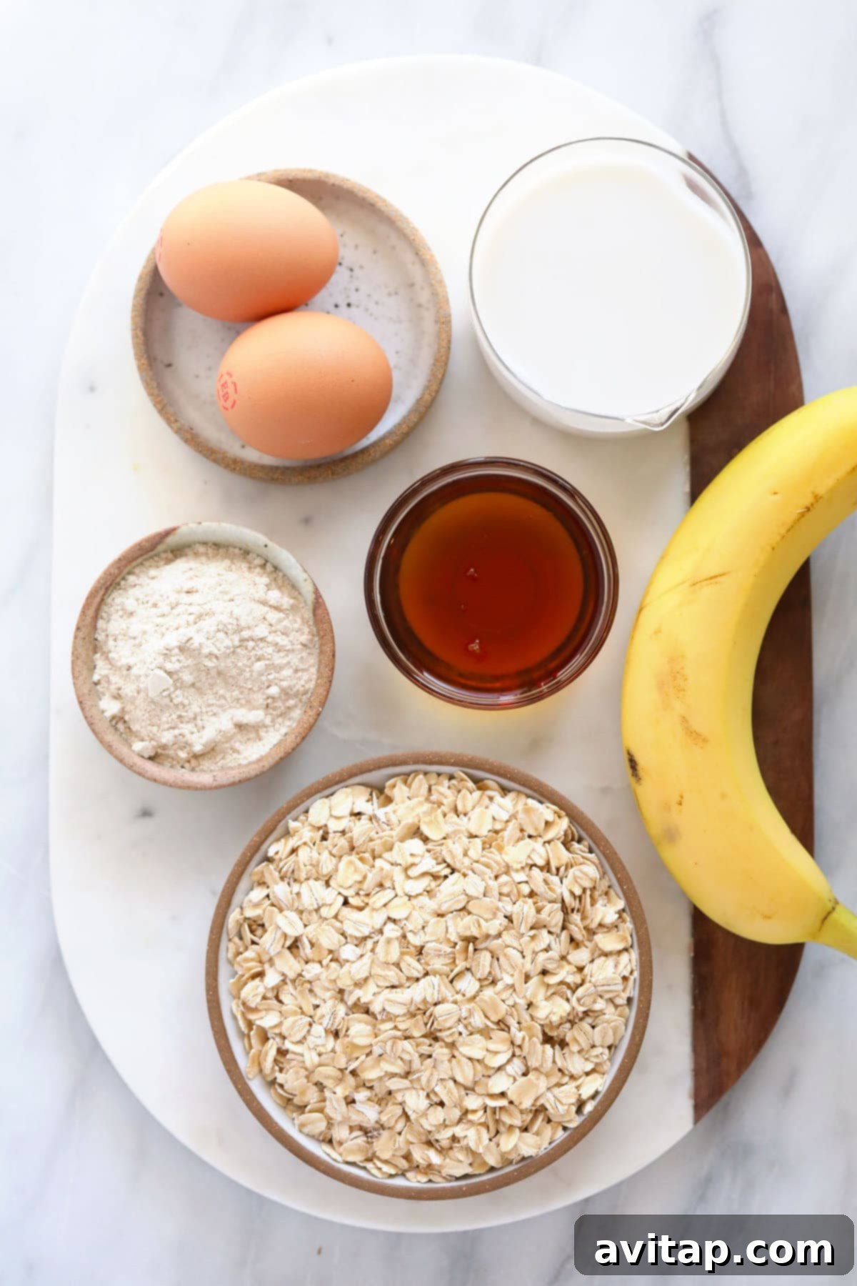 A selection of raw ingredients for baked oatmeal: oats, eggs, flour, banana, milk, and maple syrup, artfully arranged on a wooden board.