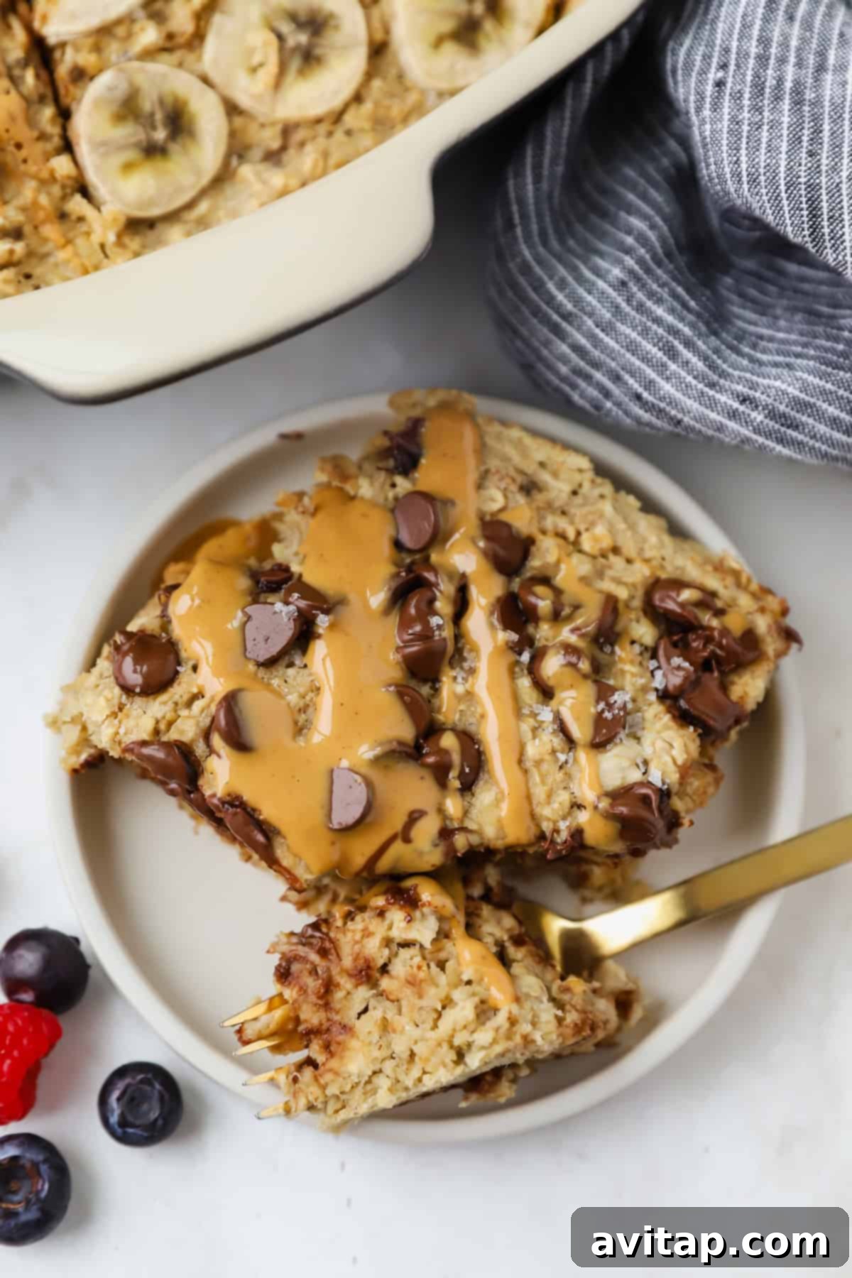 A plate with a slice of banana baked oatmeal, freshly baked, topped with chocolate chips and a peanut butter drizzle. A fork rests on the plate, accompanied by vibrant blueberries and a raspberry on the side.