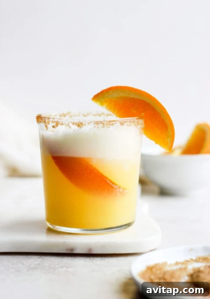 An orange cocktail garnished with a slice of orange and a frothy top.