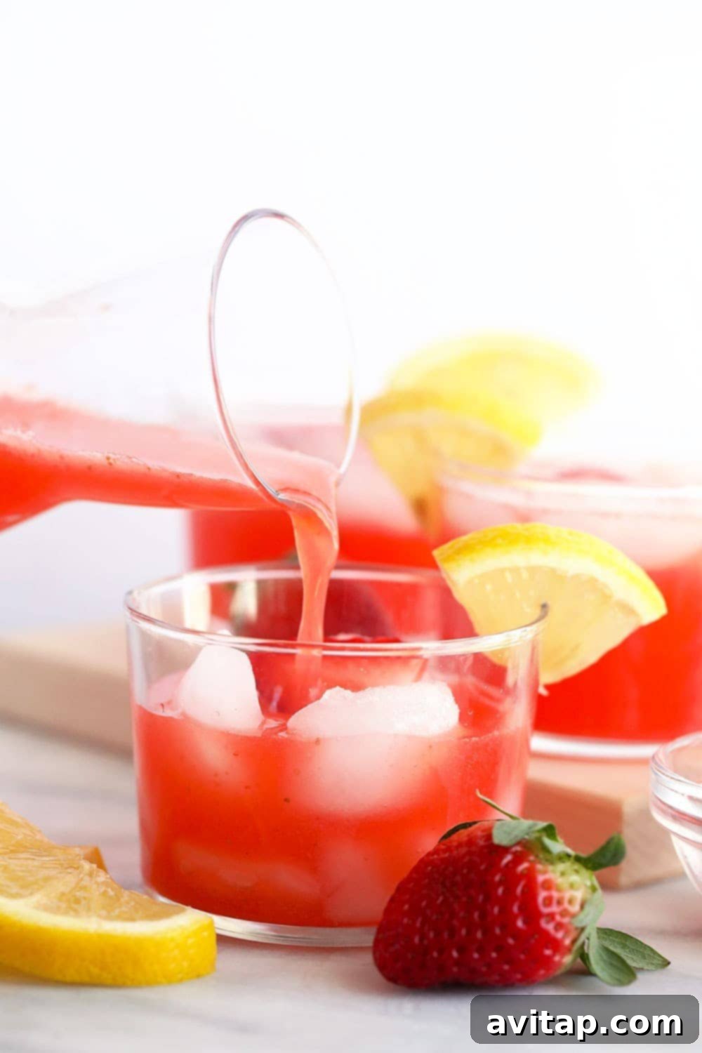 A glass of spiked strawberry lemonade being poured, garnished with a strawberry.