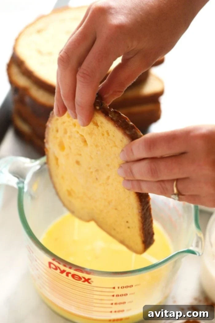 Effortless Baked French Toast 4 A slice of whole wheat bread being generously dipped into a bowl of egg mixture for making sheet pan French toast.