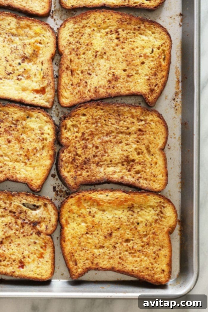 Freshly baked golden-brown French toast slices arranged neatly on a metal baking sheet, fresh out of the oven and ready for toppings.