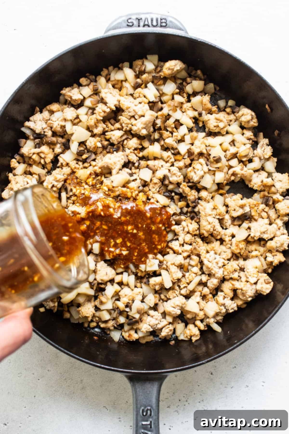 Chef pouring the homemade hoisin sauce over cooked ground chicken and vegetables in a skillet.