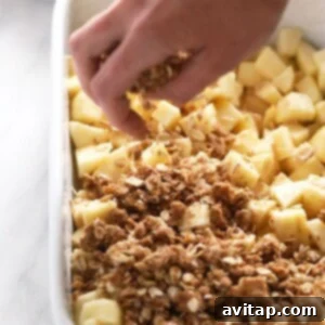 A person assembling an Apple crisp recipe, carefully spreading the oat crumble topping over the prepared apple filling in a baking dish before baking.