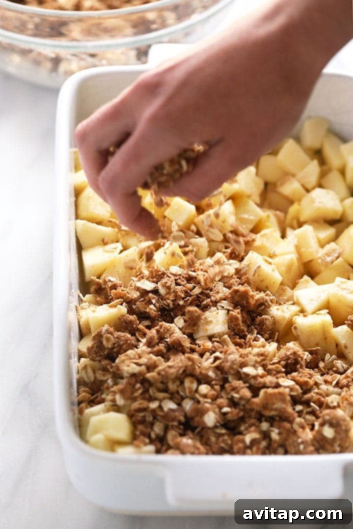 Hands sprinkling a generous amount of oat crumble topping evenly over sliced apples in a baking dish, preparing for baking.