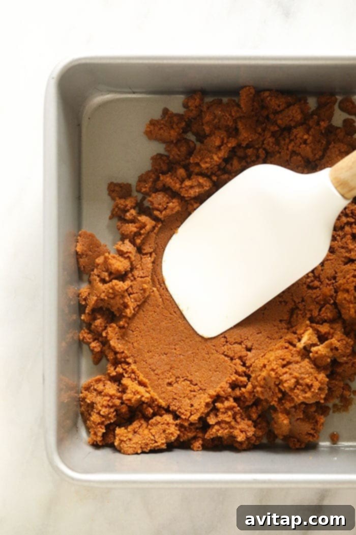 Pressing Gingersnap Crust into Pan Gingersnap crust being pressed firmly into a square baking pan, ready for the cheesecake filling.