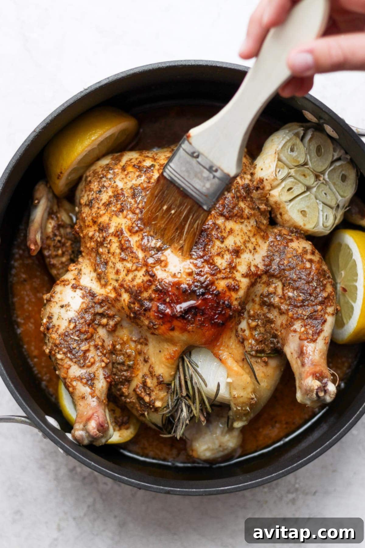 A kitchen scene showing a hand using a basting brush to coat a roasted chicken in pan juices, enhancing its flavor and moisture.