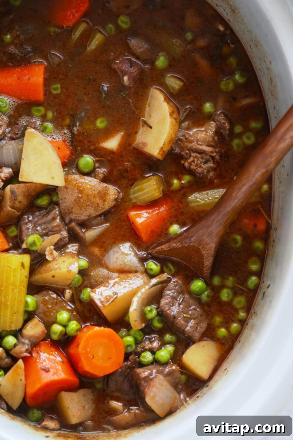 Slow cooker beef stew being gently stirred with a spoon, showcasing the rich, thick consistency and abundant ingredients.