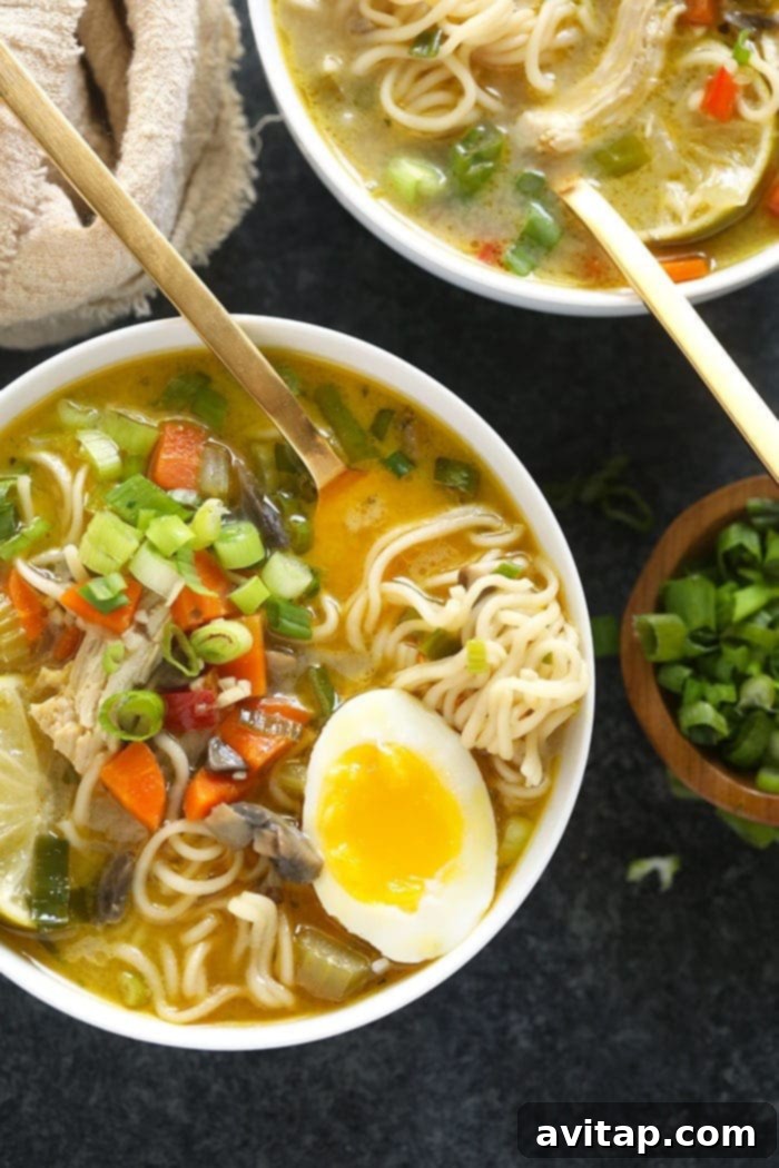 A beautifully presented, deep bowl of Asian Chicken Noodle Soup with ramen noodles, garnished exquisitely with vibrant green onions, a wedge of fresh lime, and a perfectly cooked soft-boiled egg, invitingly ready to be savored.