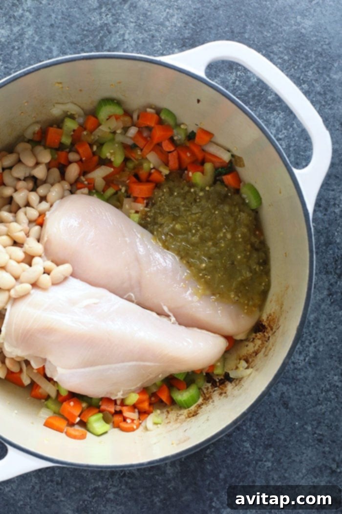 Array of fresh ingredients including carrots, celery, and a jar of salsa verde laid out around a dutch oven, ready for Green Monster Spicy Chicken Noodle Soup preparation.