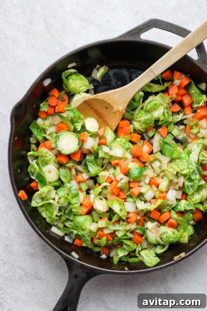 A large skillet filled with vibrant, sautéed vegetables, including carrots, celery, onions, and Brussels sprouts, for cornbread stuffing.