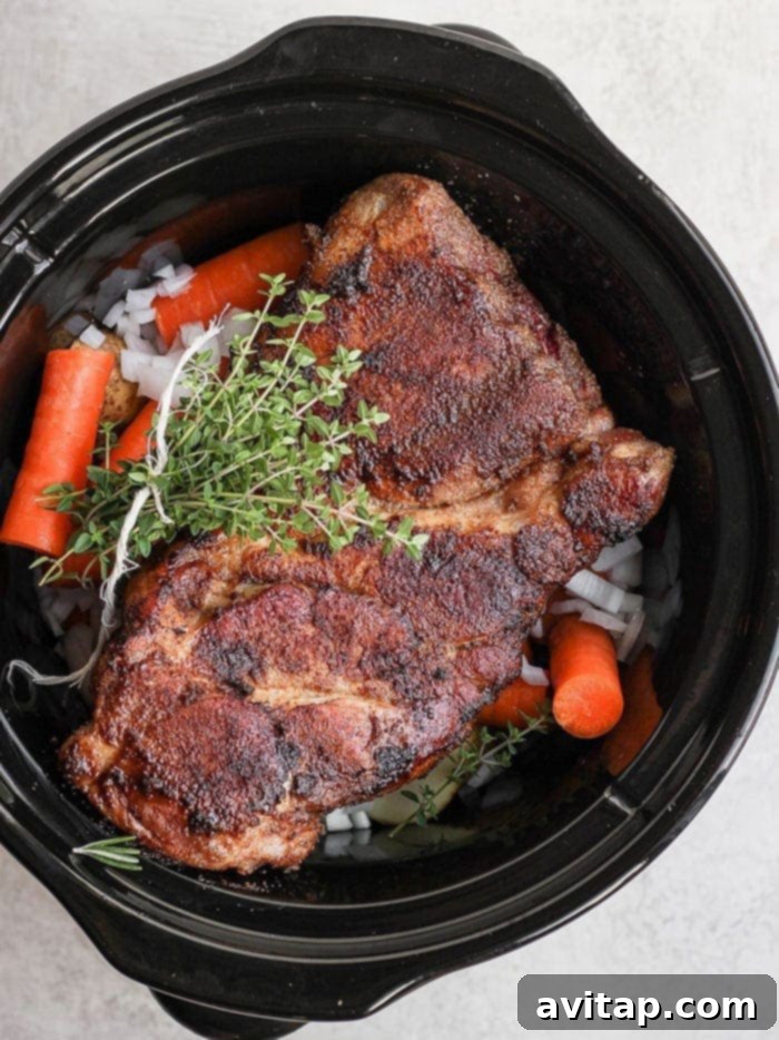 A seasoned pork shoulder settling into the slow cooker on a bed of vegetables.