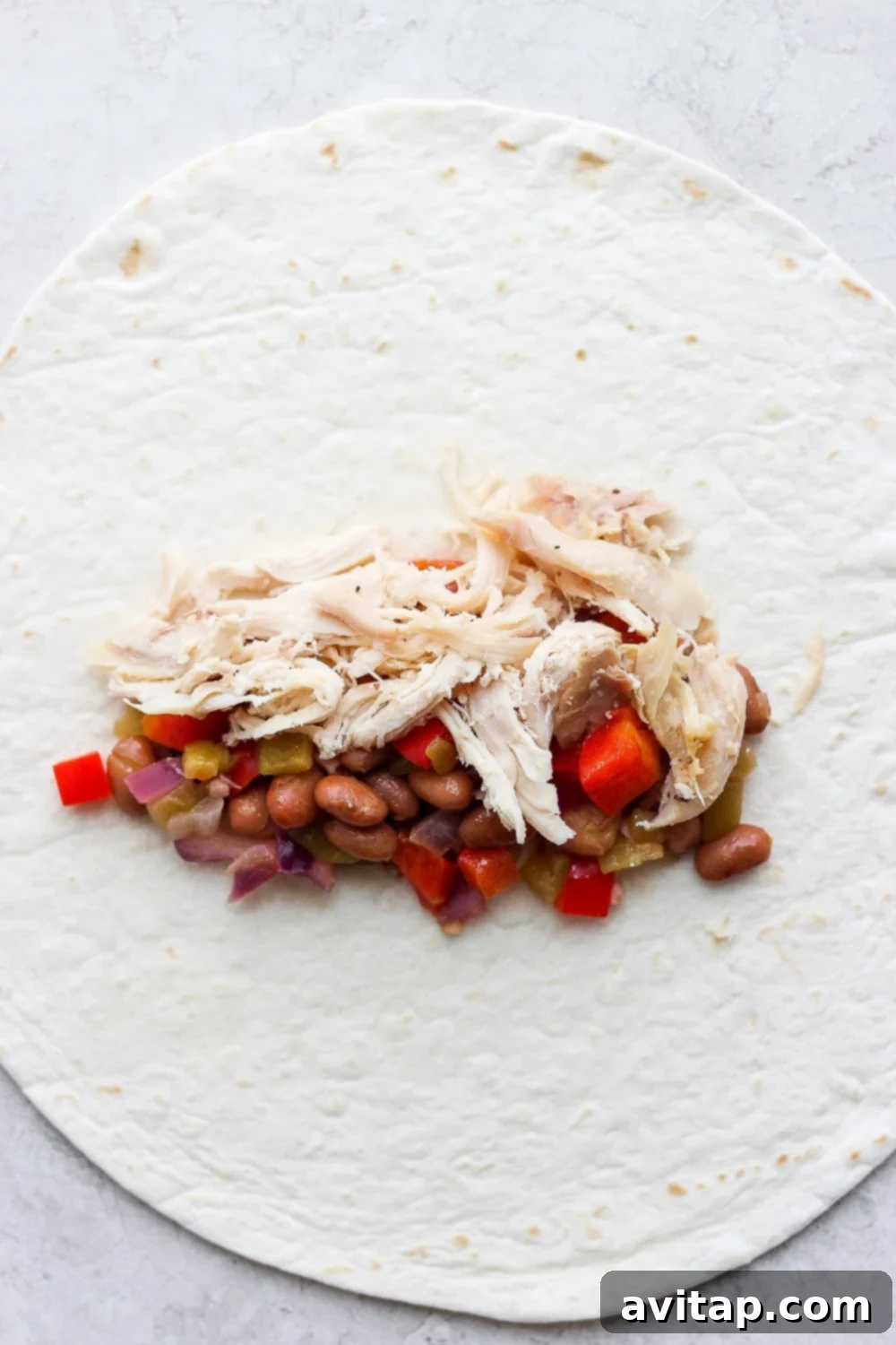 Hands carefully rolling a flour tortilla filled with shredded chicken and vegetable mixture, preparing it for baking.