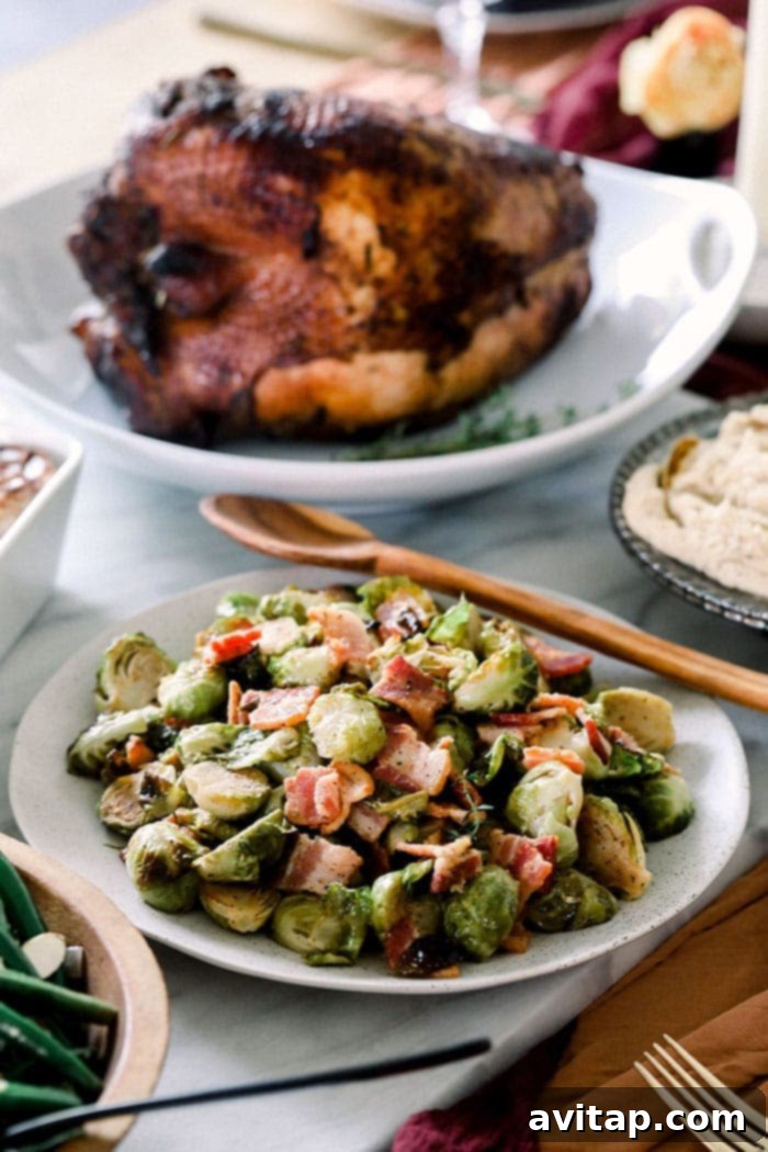 A bowl of roasted Brussels sprouts with bacon on a festive holiday table with a roasted turkey in the background, set for a meal.