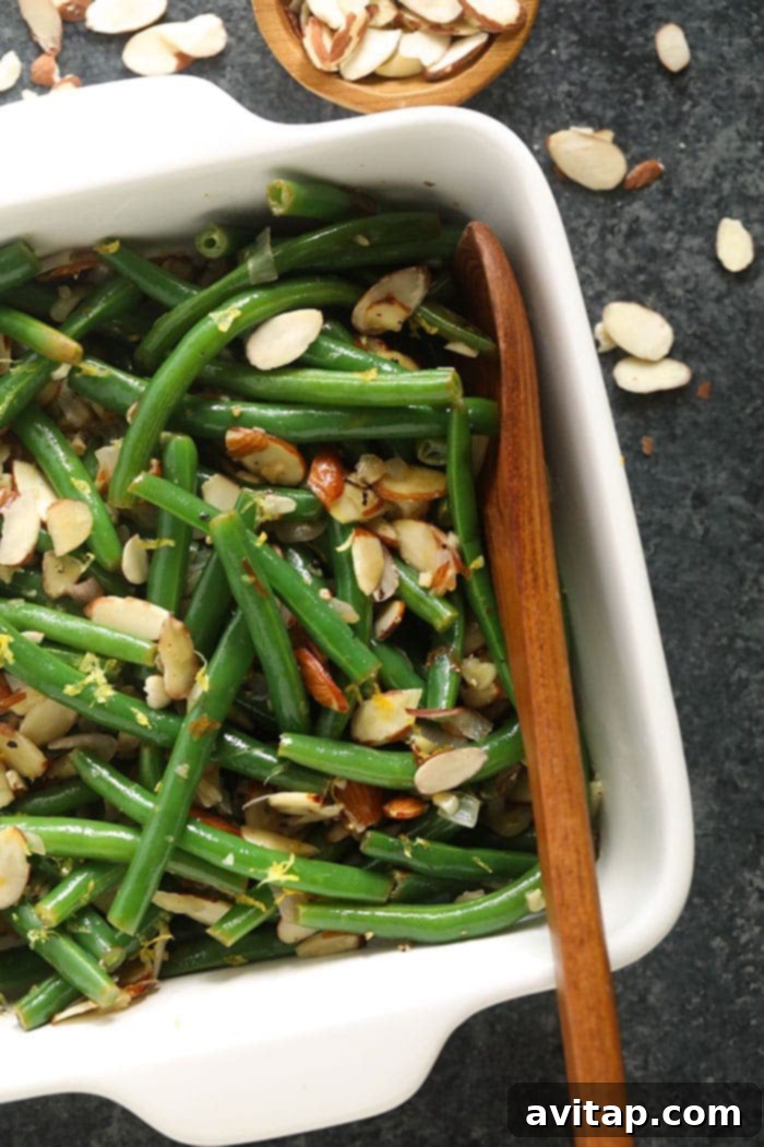 Green Beans Almondine is a fresh, buttery holiday side made with perfectly blanched green beans, a delicious slivered almond topping, and fresh lemon zest! Green Beans Almondine served in a beautiful white casserole dish, garnished with fresh lemon zest and parsley.