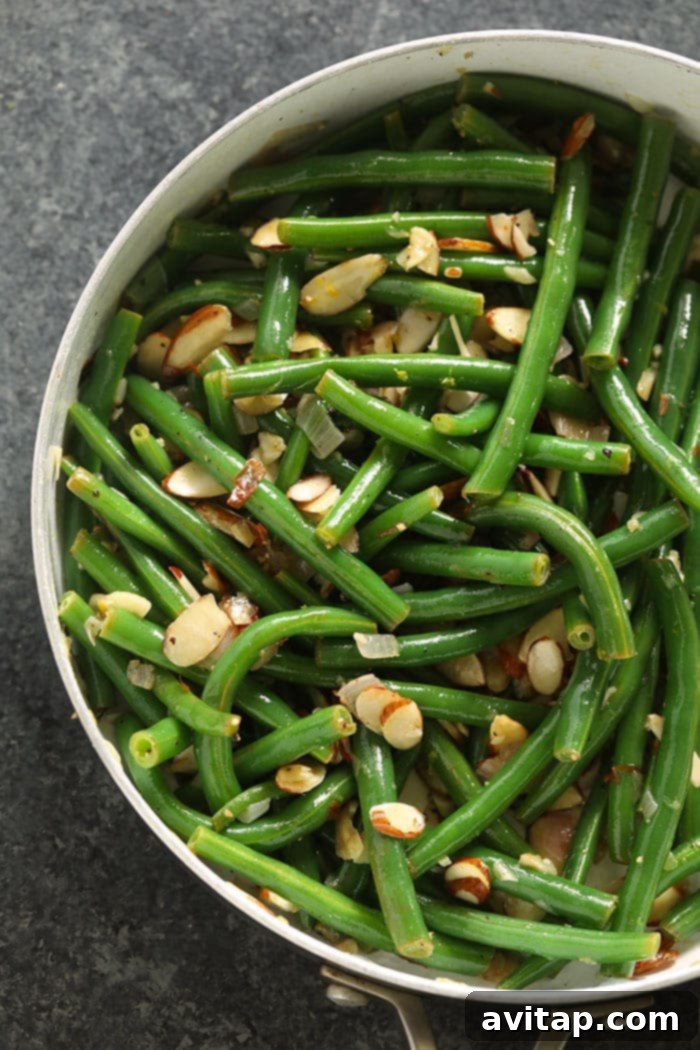 Our Green Beans Almondine recipe is crunchy, buttery, and has a delightful hint of lemon! Green Beans Almondine being sautéed in a large non-stick pan, with butter, garlic, and golden almonds.