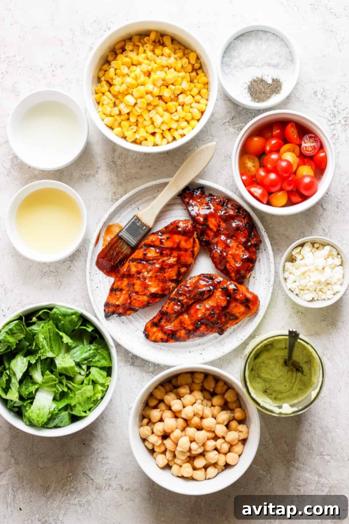 A collection of fresh BBQ chicken salad ingredients laid out on a table, including romaine, corn, tomatoes, garbanzo beans, and feta cheese.