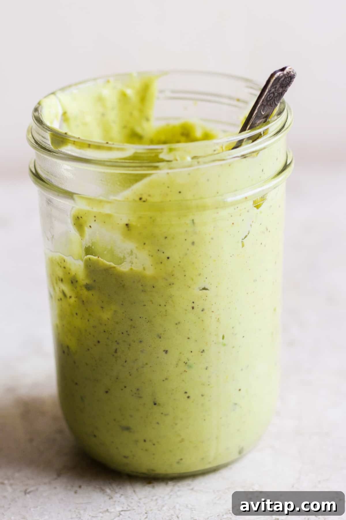 A close-up of a jar filled with creamy green avocado ranch dressing, with a spoon resting in it.