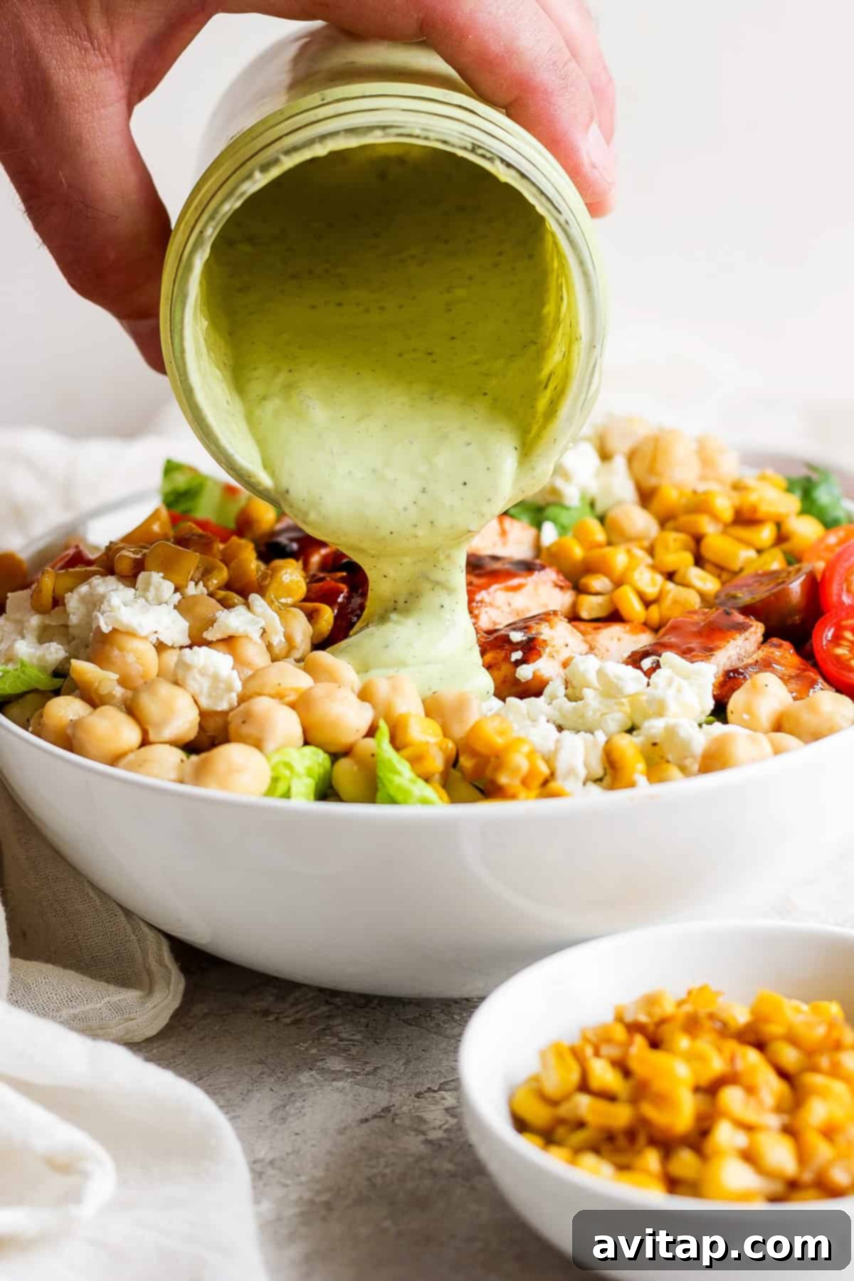 A person pouring avocado ranch dressing from a small jar over a large bowl of BBQ chicken salad.