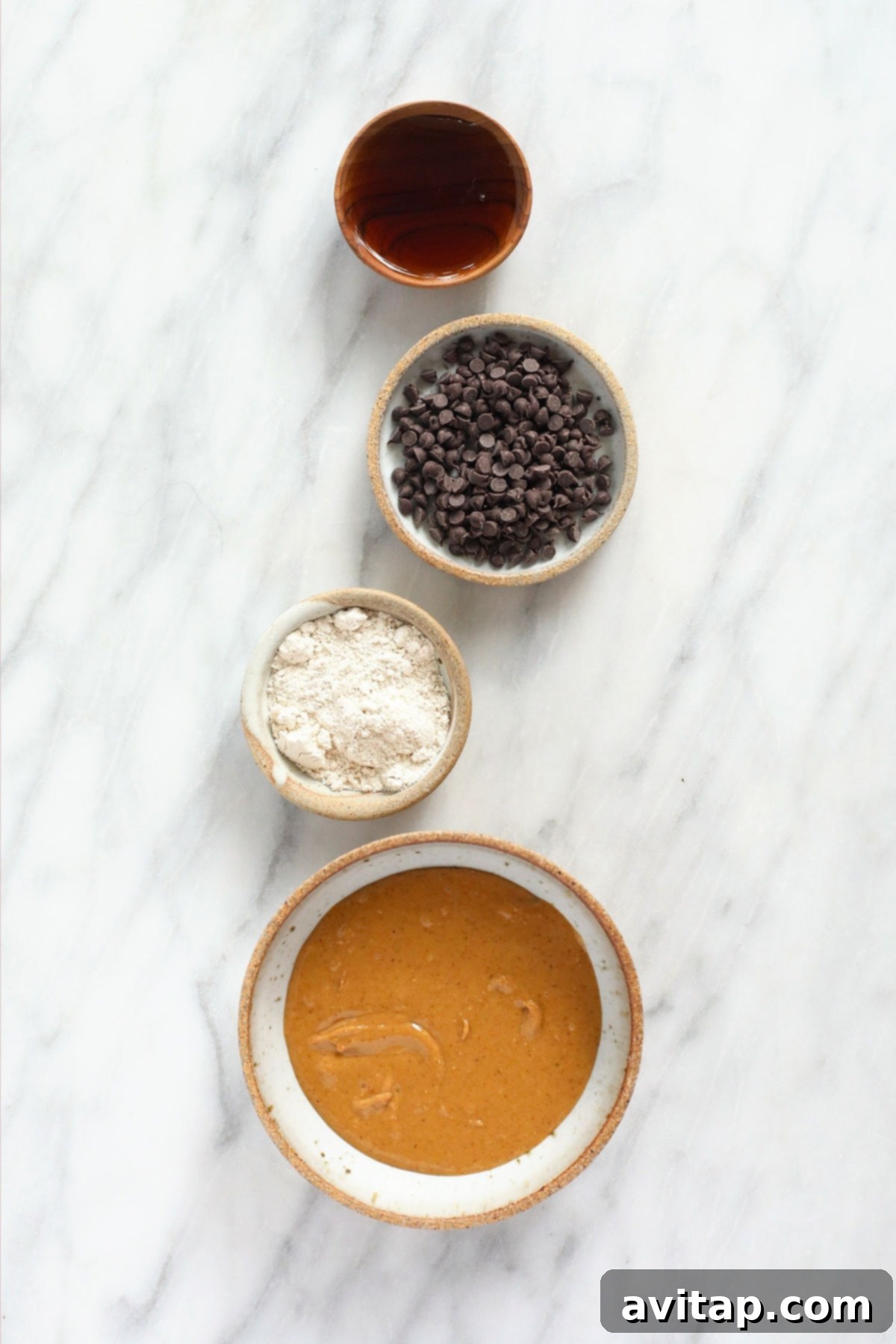 Edible Vegan Cookie Dough Delights 3 A vibrant selection of essential ingredients for vegan cookie dough bites: maple syrup, dairy-free chocolate chips, ground oat flour, and creamy peanut butter, artfully arranged in small bowls.