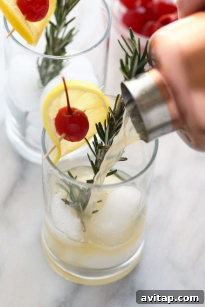 Hand pouring a Rosemary Collins cocktail into a tall glass filled with ice, garnished with rosemary and lemon.