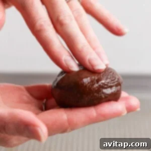 A person shaping a ball of dark brown cookie dough in their hands, demonstrating the molding process for the protein cookies.