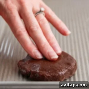 A hand presses down on a round chocolate cookie dough piece on a baking sheet, illustrating the process of flattening and adding toppings.
