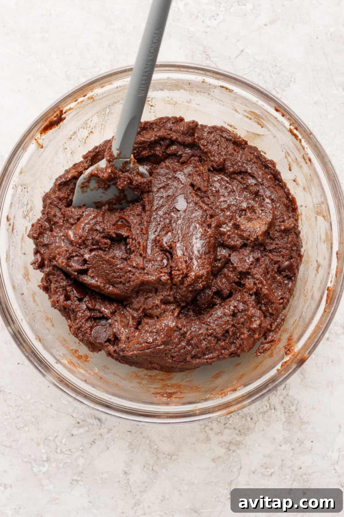 A glass bowl filled with rich, thick chocolate cookie dough, ready for shaping, with a gray spatula resting inside.