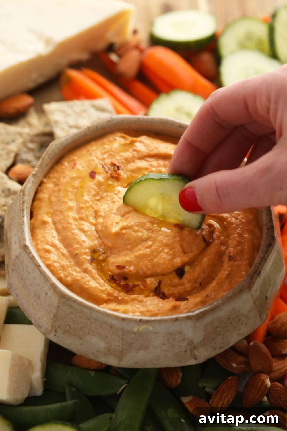 A close-up of creamy roasted red pepper hummus in a bowl, with a sliced cucumber being dipped into it, highlighting its rich texture and vibrant color.