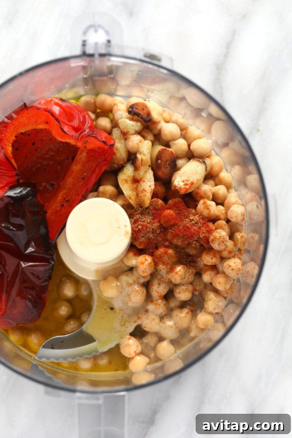 All the vibrant ingredients for roasted red pepper hummus, including chickpeas, tahini, lemon, spices, and roasted peppers with garlic, are placed in a food processor bowl, ready for blending.