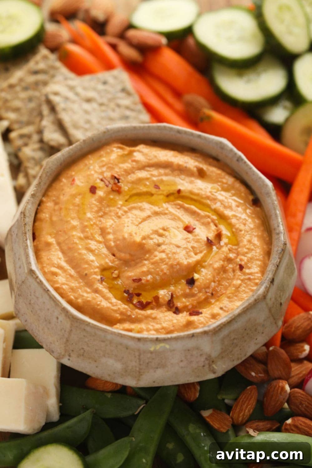 A beautifully presented bowl of homemade roasted red pepper hummus, surrounded by a colorful array of dipping options including crackers, various sliced fresh vegetables, and warm pita bread.
