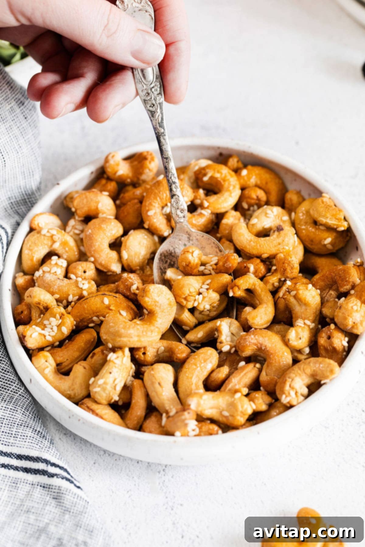 A bowl filled with golden brown roasted cashews, garnished with sesame seeds and a spoon resting inside, highlighting their delicious crunch.