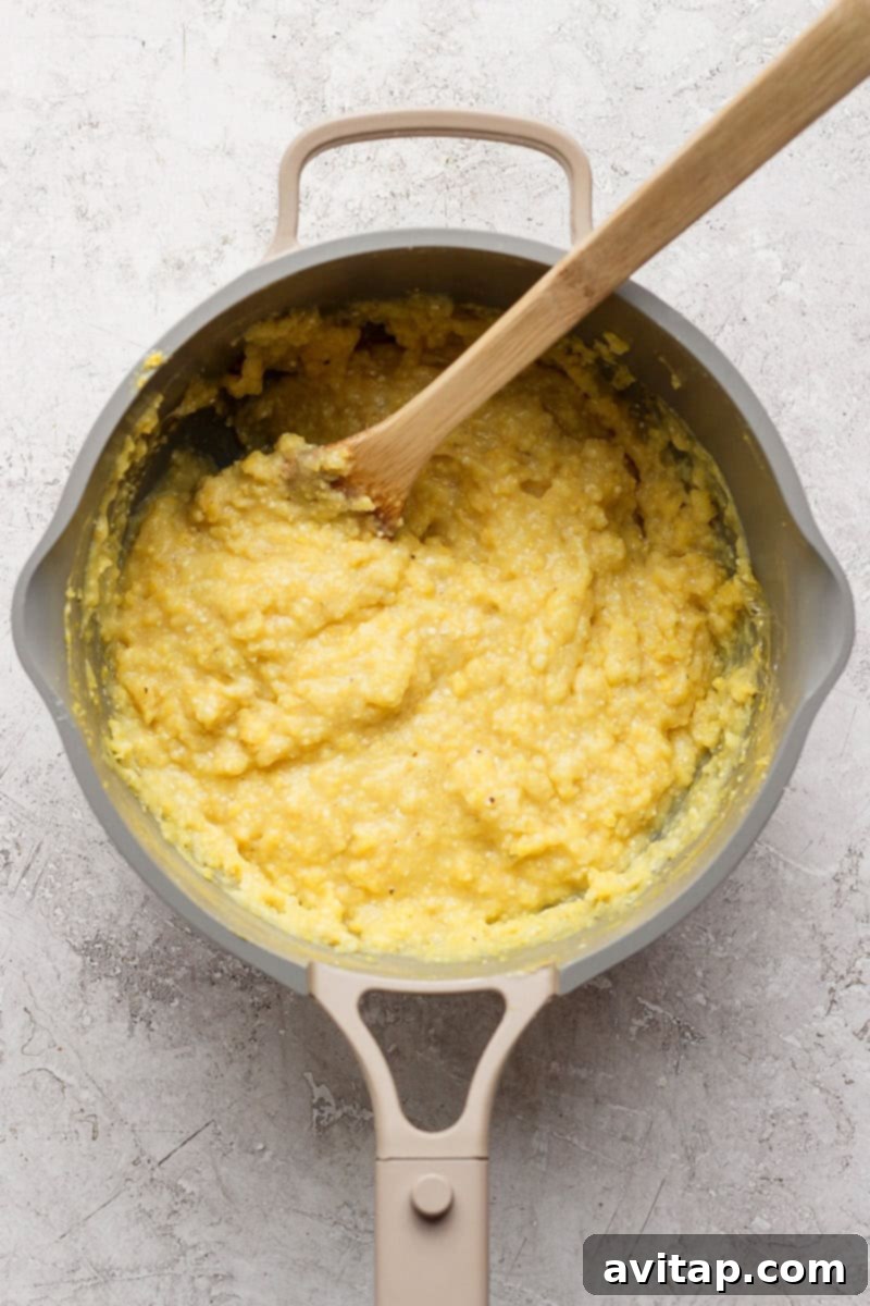 A pot on a light surface contains thick yellow grits being stirred with a wooden spoon.