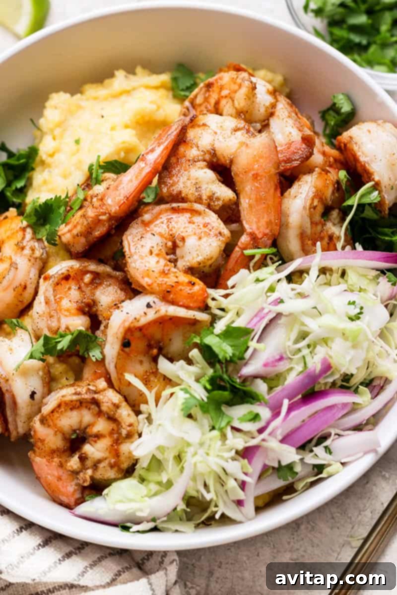 A bowl of seasoned shrimp atop creamy polenta, garnished with fresh herbs, sliced cabbage, red onion, and cilantro. A lime wedge is partially visible on the side.