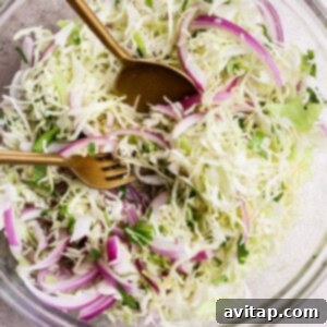 A hand mixing coleslaw in a clear glass bowl with a fork and spoon. The coleslaw contains shredded cabbage, red onions, and herbs.