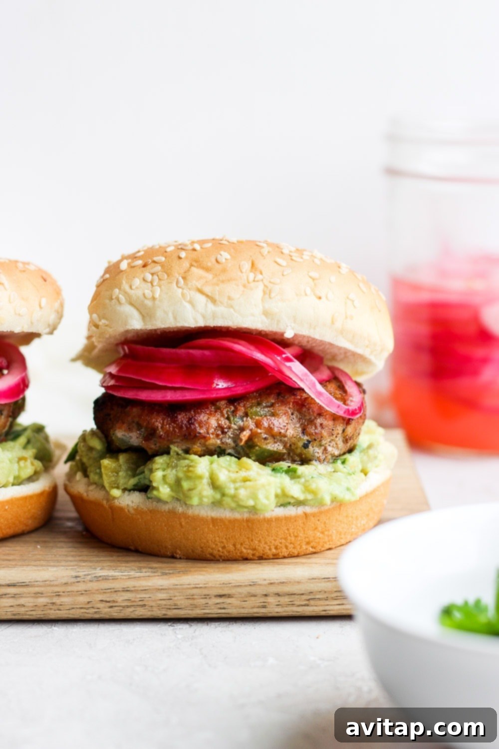 Say hello to the Best Juicy Turkey Burger you've ever had! These turkey burgers are always delicious, and never dry. Enjoy! Two juicy turkey burgers on buns, topped with vibrant purple onions and avocado mash, ready to be served on a wooden board.