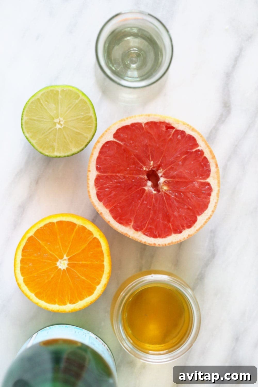 Essential ingredients for homemade hard seltzer laid out: fresh citrus fruits, various herbs, a bottle of vodka, and sparkling water.