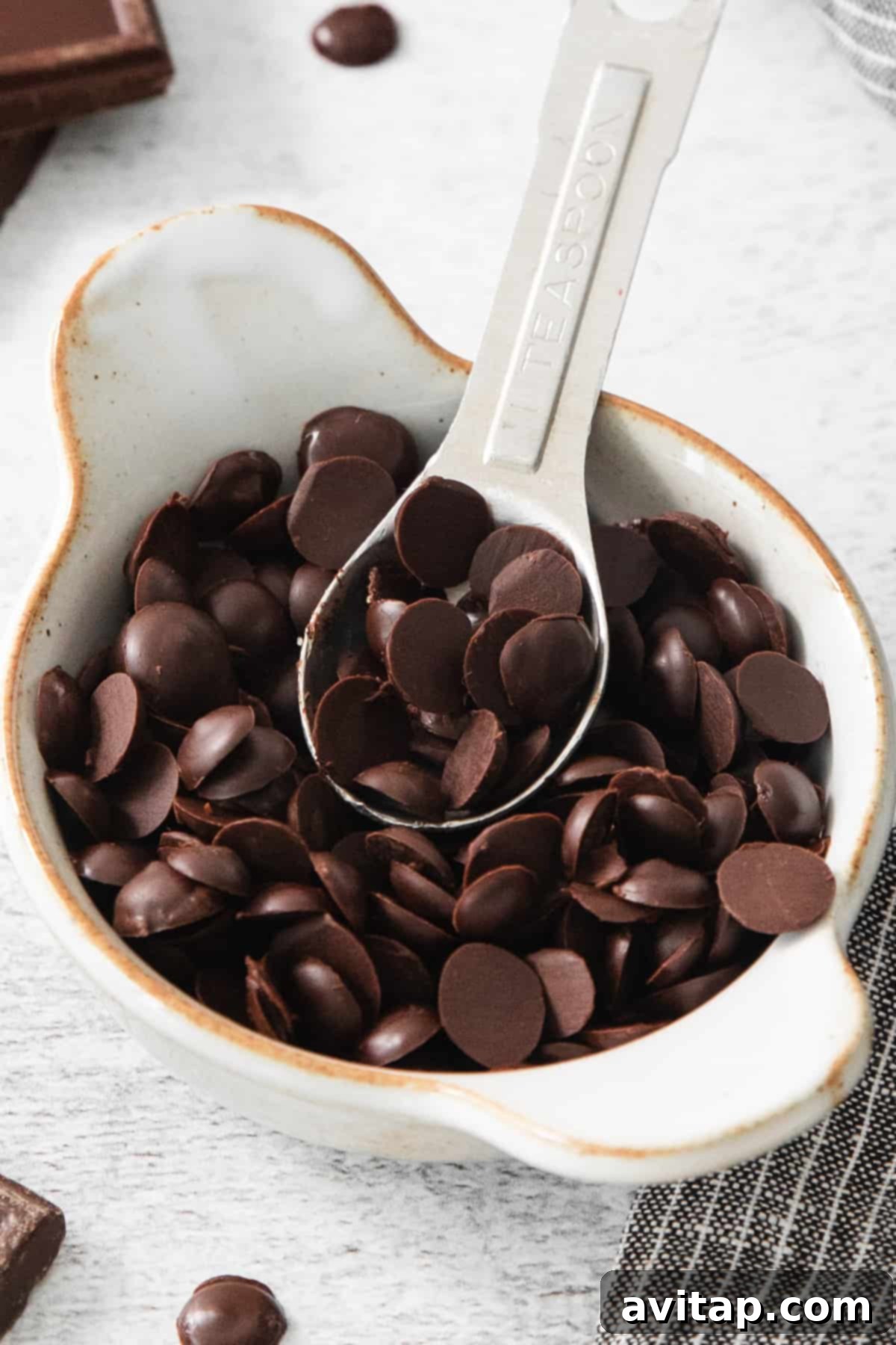 A white bowl filled with freshly made homemade vegan chocolate chips, with a small silver spoon resting inside, inviting a taste.