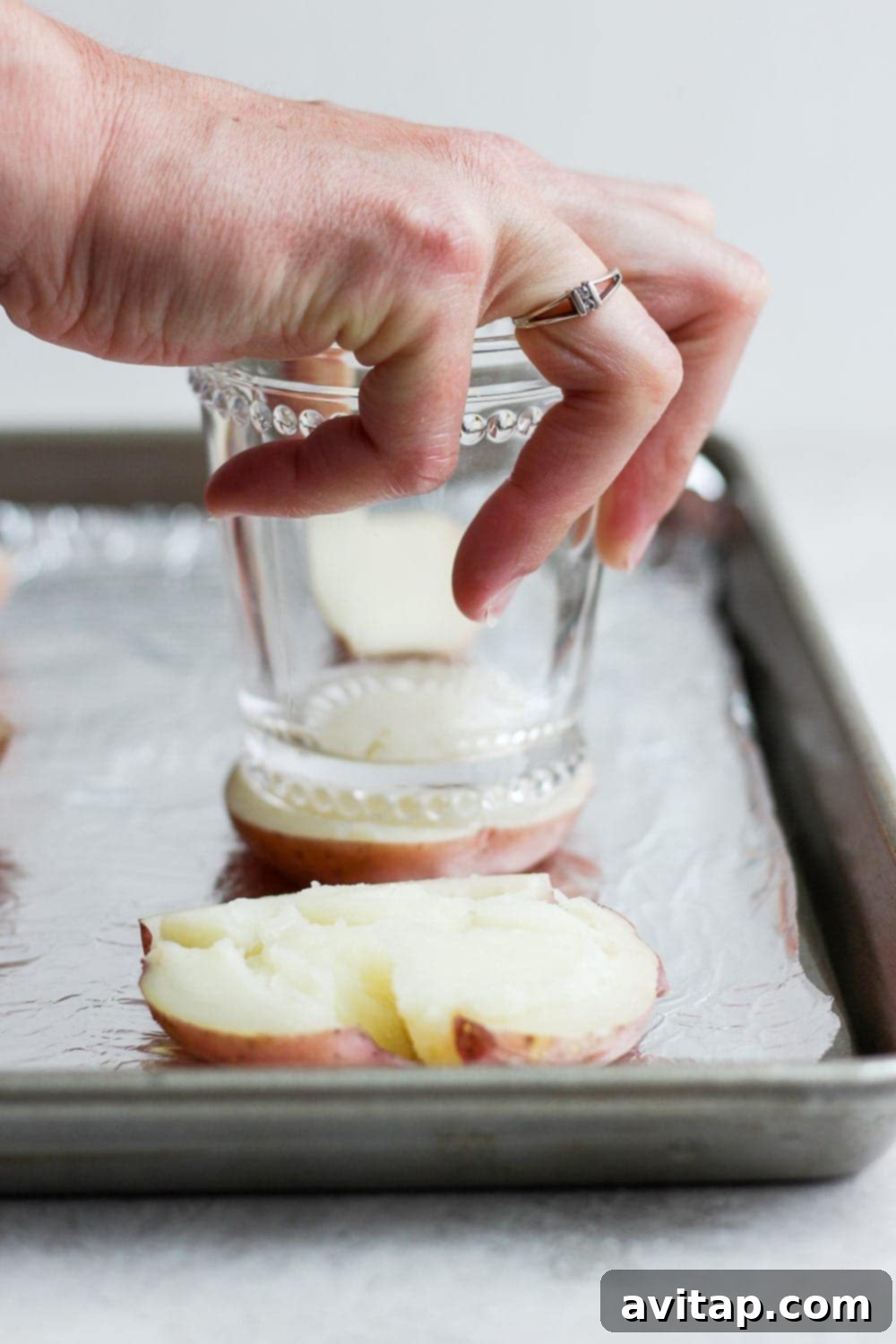 Achieving the ideal smashed potato shape with a flat-bottomed tool. A boiled red potato half being smashed flat with the bottom of a clear drinking glass.