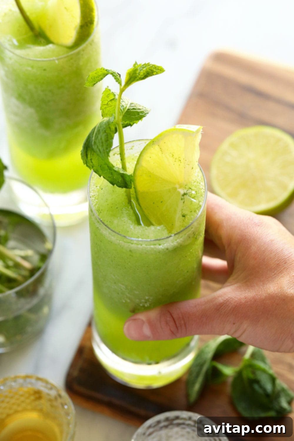 Hand holding a tall glass of vibrant green frozen mojito, garnished with fresh mint and a lime wedge, against a bright background.
