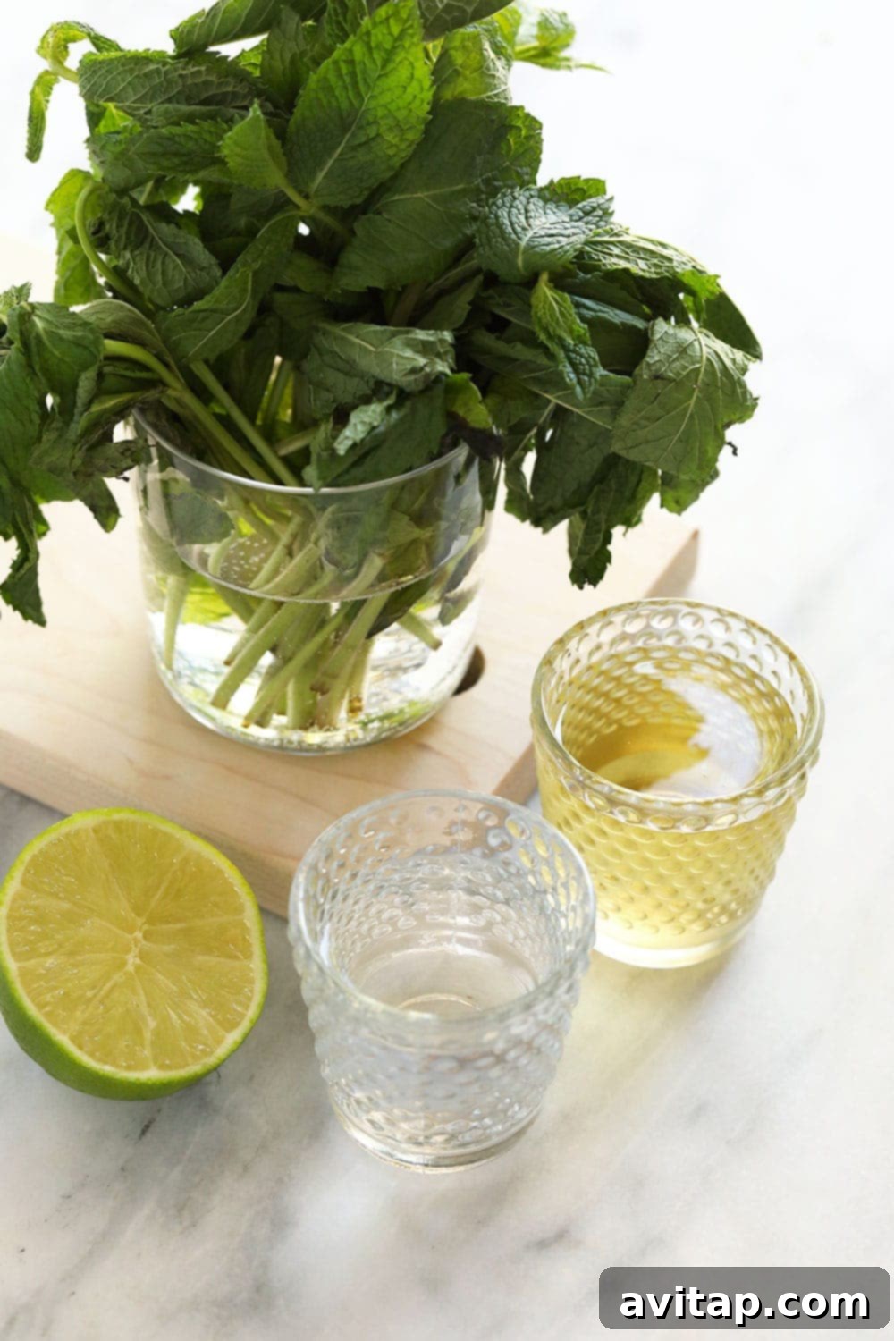 Fresh ingredients laid out for frozen mojitos: a glass with fresh mint, a small pitcher of simple syrup, a bottle of white rum, and a bowl of fresh limes.