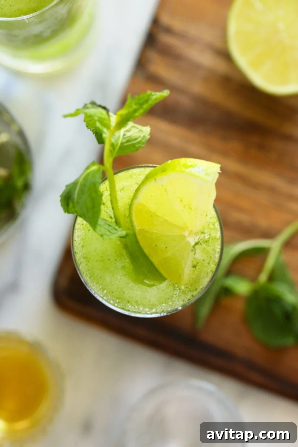 Top-down view of a vibrant green frozen mojito in a clear glass, garnished with fresh mint sprigs and a lime wheel, sitting on a wooden surface.