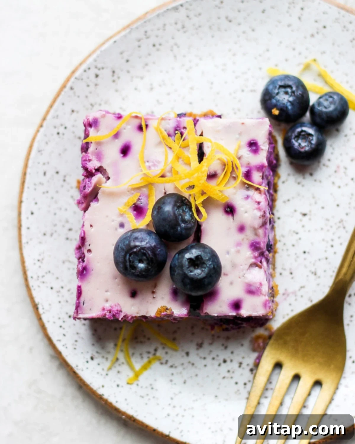 Enjoy these vibrant Lemon Blueberry Cheesecake Bars – a perfect blend of tart and sweet with a classic graham cracker crust. A delectable lemon blueberry cheesecake bar on a white plate, garnished with fresh blueberries and lemon zest.