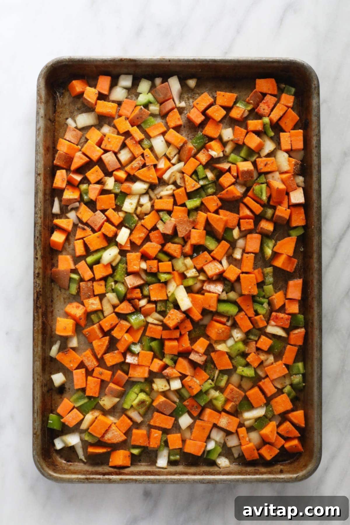 Raw sweet potato, bell pepper, and onion diced and seasoned on a baking pan before roasting.