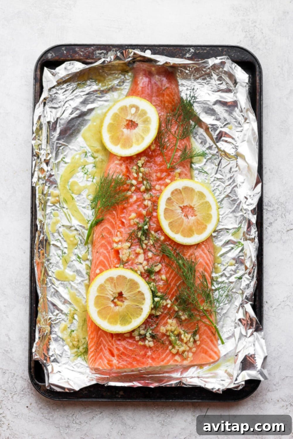 Large salmon fillet beautifully arranged on tin foil with fresh lemon slices, dill, and garlic, showcasing the aromatic marinade before baking.