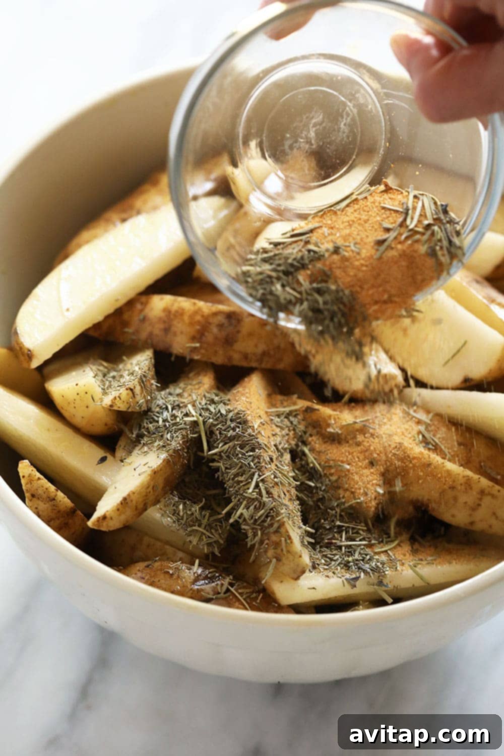 Pouring herbs and spices onto potato wedges in a bowl before baking.