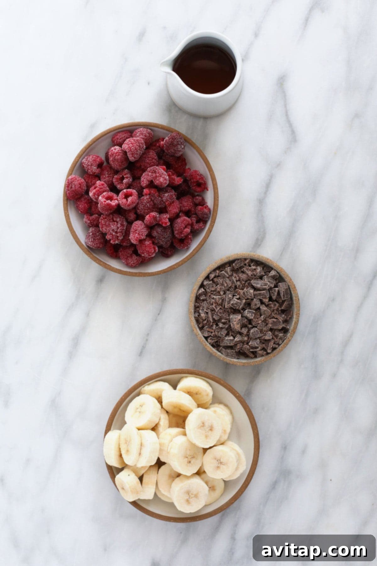 The essential ingredients for Raspberry Chocolate Chunk Nice Cream laid out on a surface, including frozen bananas, frozen raspberries, almond milk, vanilla extract, and chocolate chunks, ready for blending.