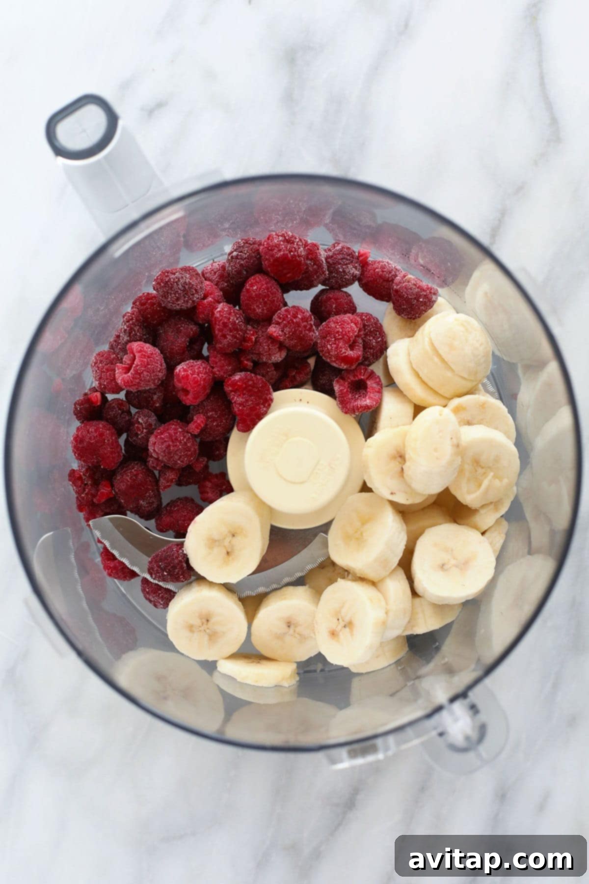 A high-angle shot of frozen bananas and raspberries, along with a splash of almond milk and a hint of vanilla extract, being blended to perfection in a food processor for a creamy nice cream base.