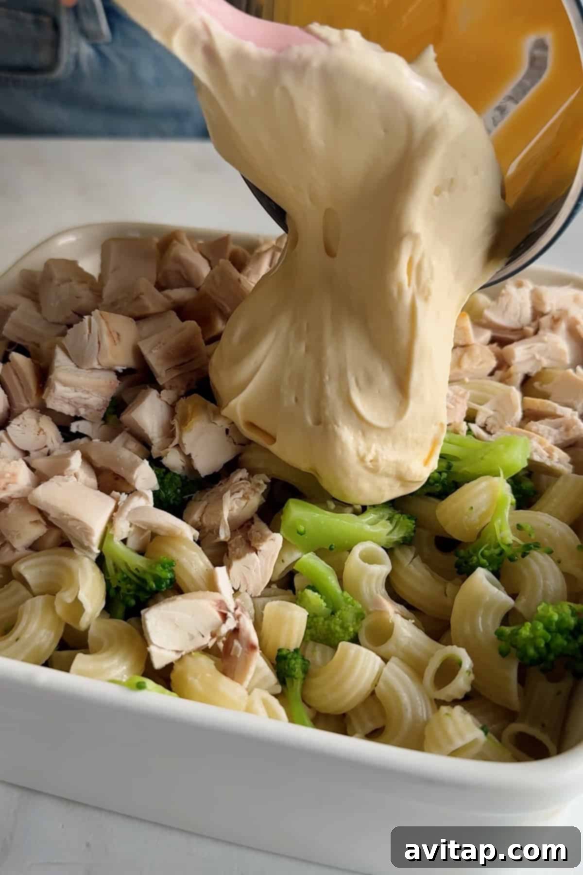 Cheese sauce being poured over pasta, chicken, and broccoli in a baking dish.