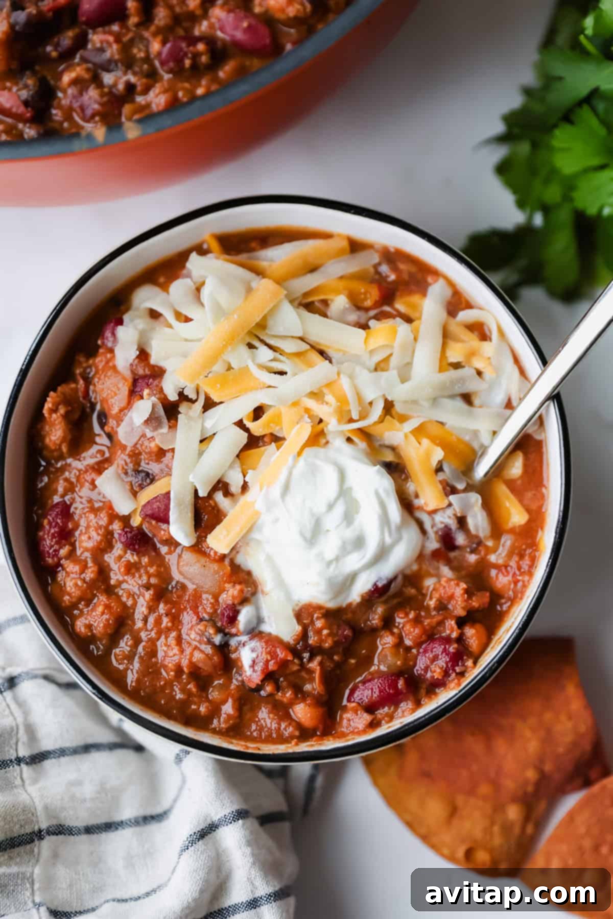 A bowl of chili topped with shredded cheese and a dollop of sour cream, with a spoon, tortilla chips, and a striped napkin beside it.